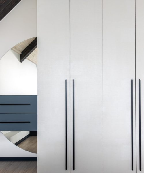 White fronts with long black handles and a round mirror reflect blue floating drawers, wooden beams and soft light shape a neat space with scandi and japandi feel.